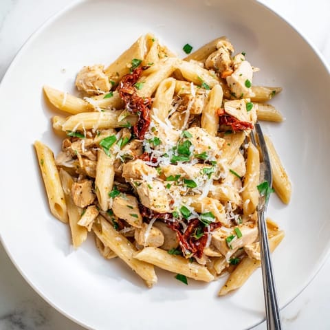 A close-up of Creamy Sun-Dried Tomato Chicken Pasta reveals glossy sauce clinging to penne, golden chicken, and vibrant sun-dried tomato ribbons.  